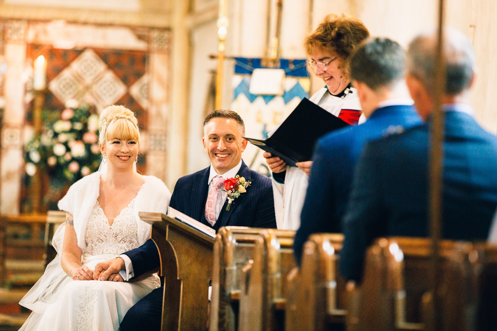 Bride and groom seated and smiling during the ceremony, the vicar behind