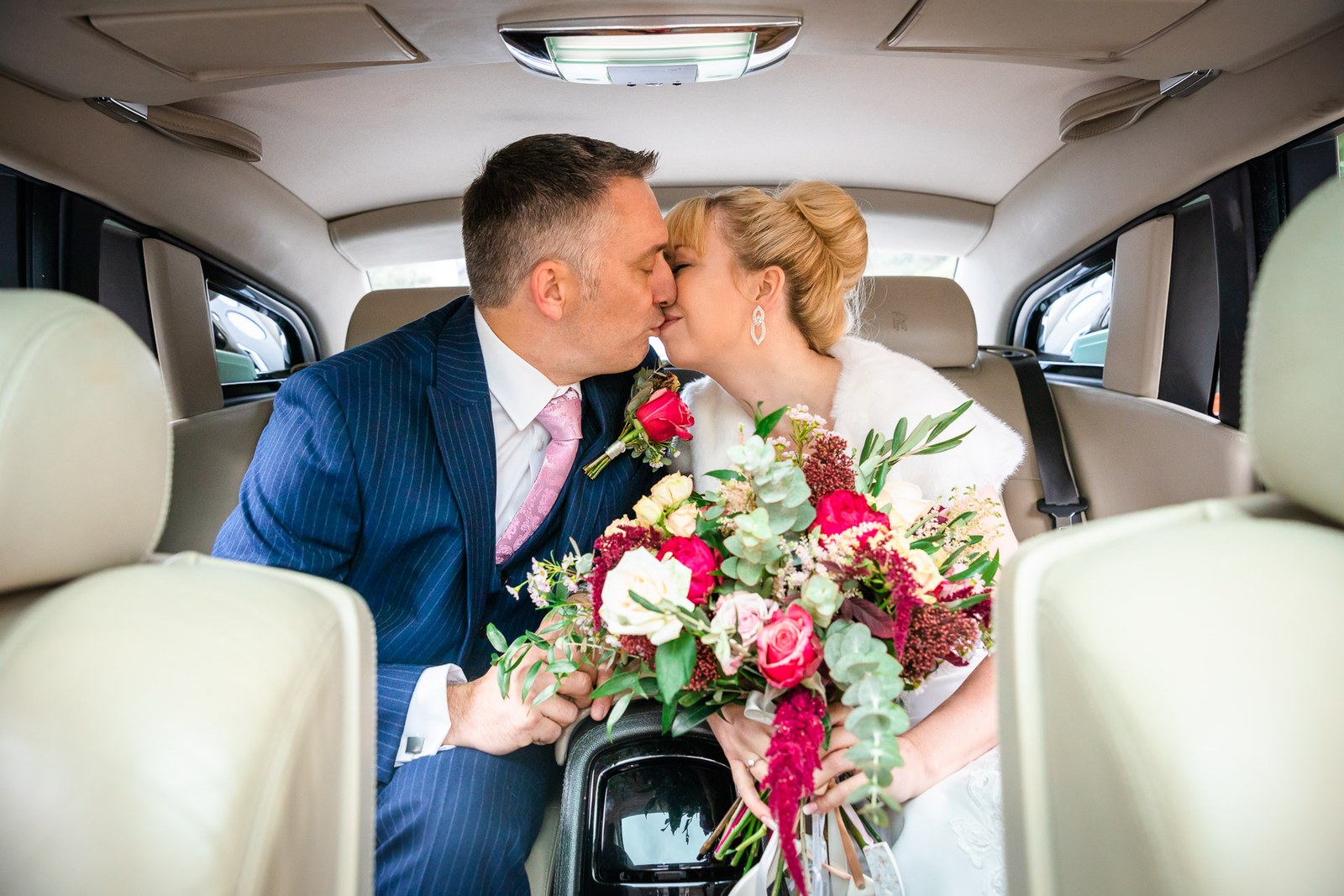 Bride and groom kissing in the back of the wedding car, red bouquet in hand