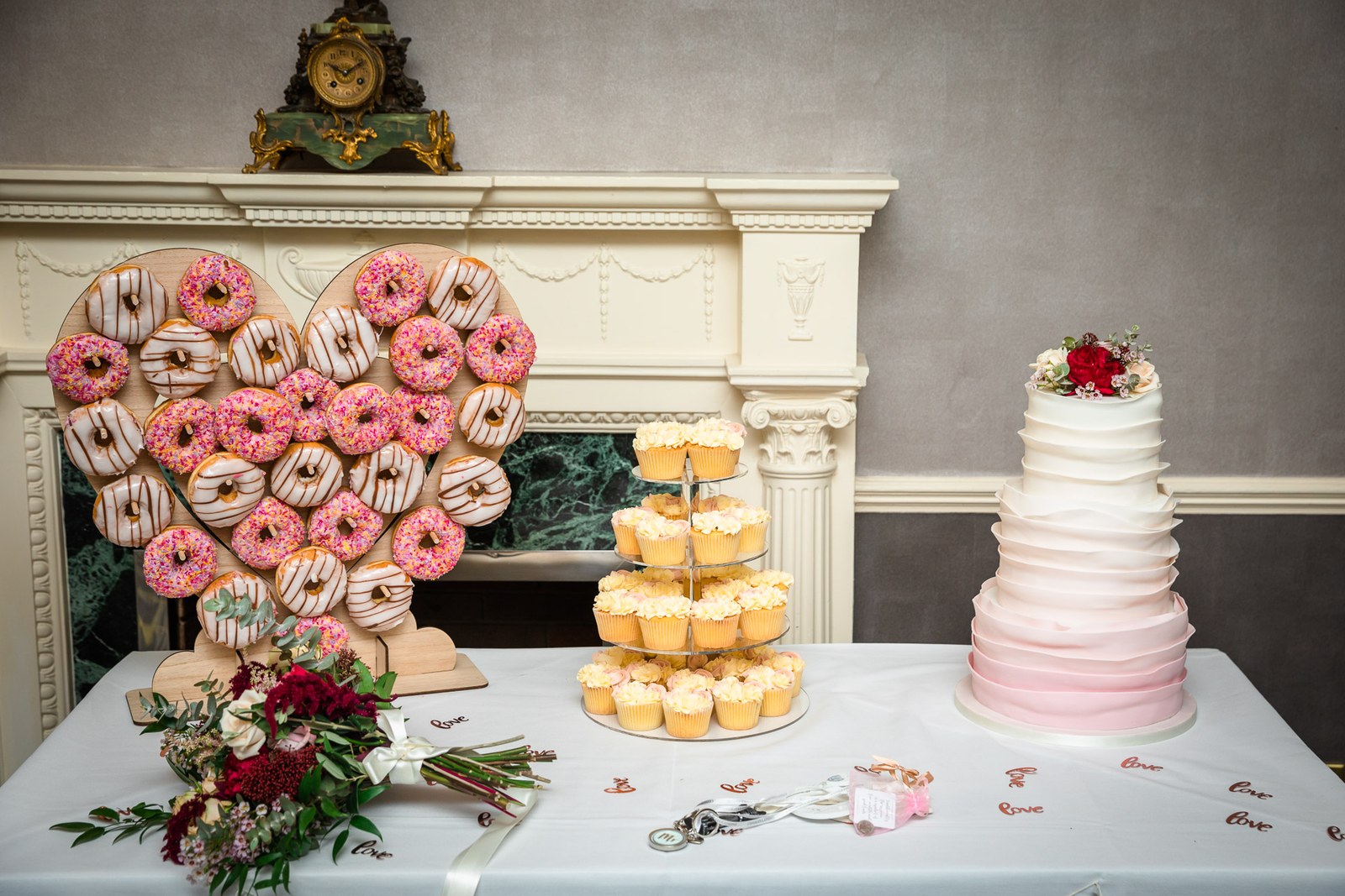 Dessert table with a heart-shaped doughnut wall, cupcake tower and ombre cake by a fireplace