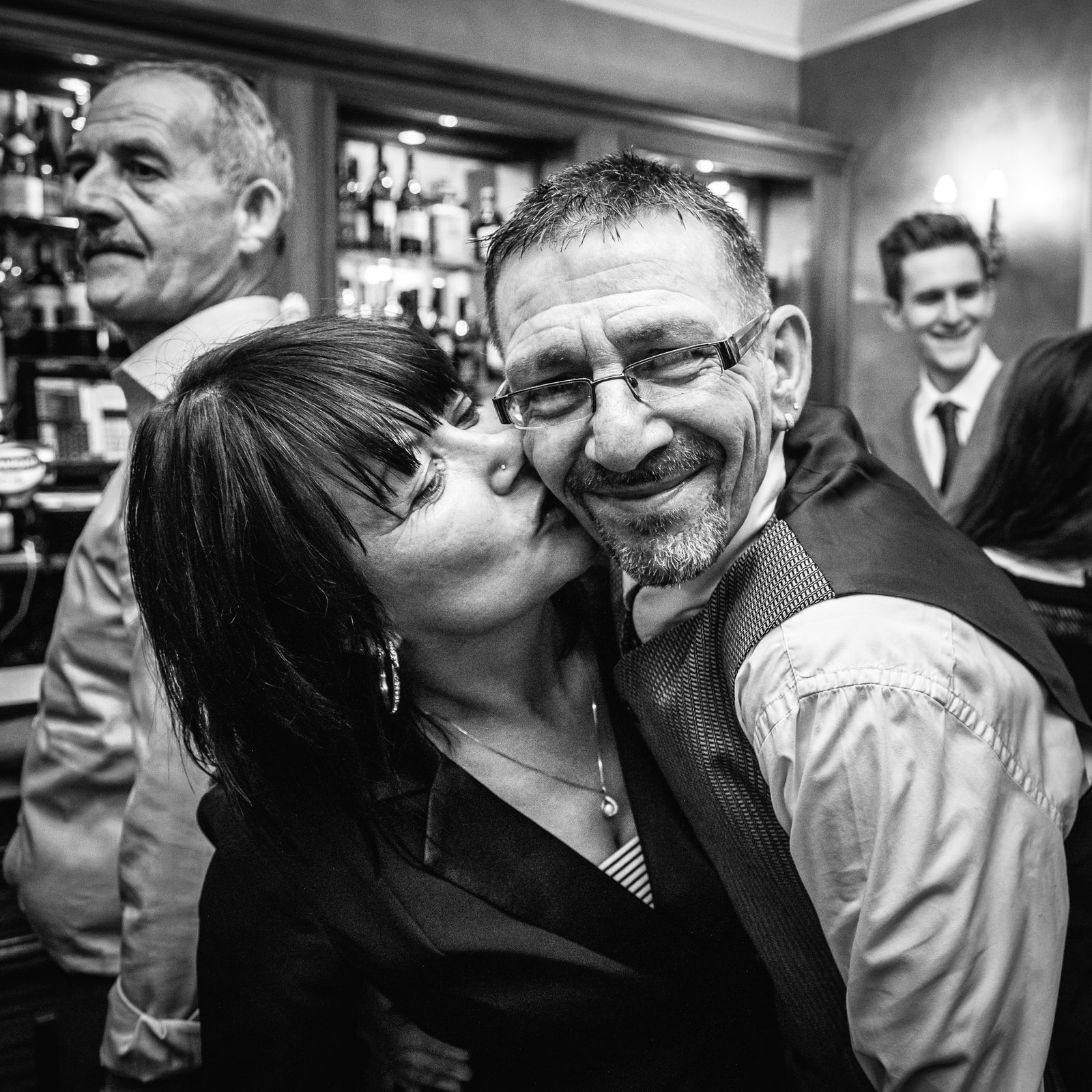 Black and white of a guest kissing a smiling man on the cheek at the bar