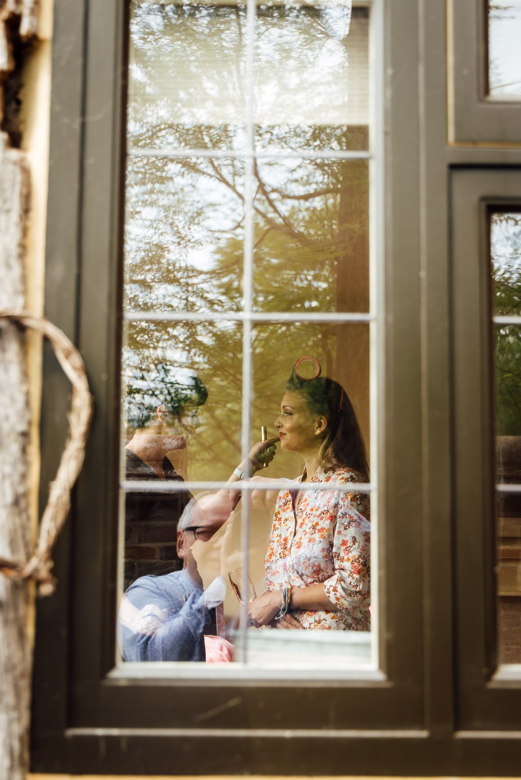 Bride having her make-up done, seen through a manor window with trees reflected in the glass