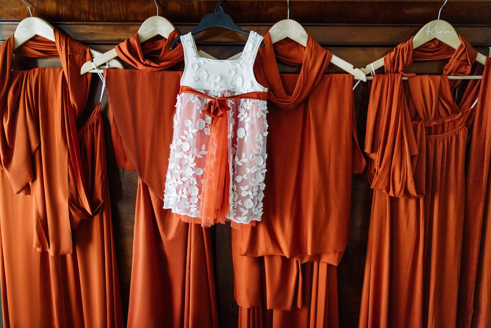 Rust-orange bridesmaid dresses and a floral flower-girl dress hanging from a beam