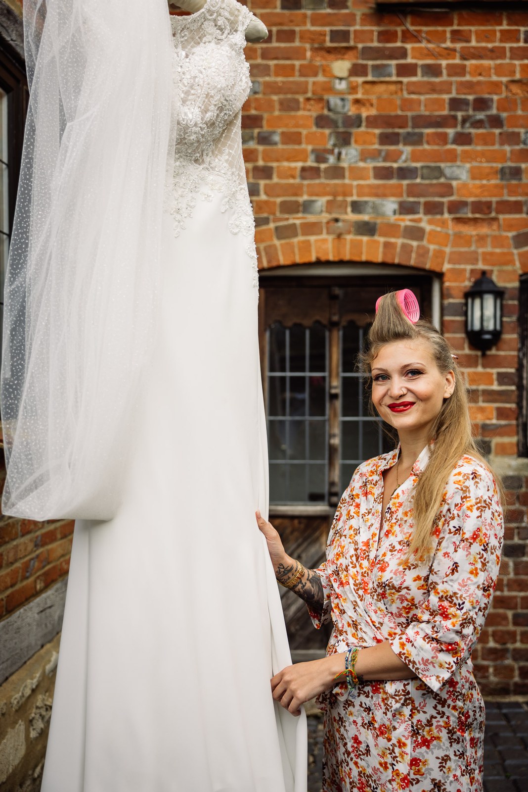 Bride in a floral robe beside her wedding gown hung against a red-brick wall