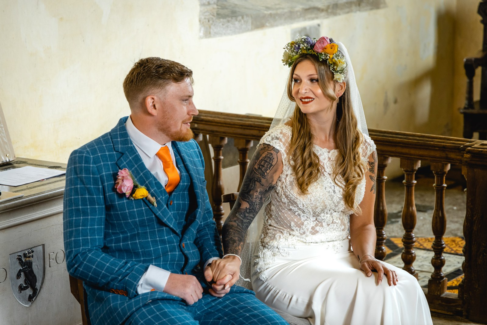 Bride and groom seated during the ceremony beside a carved heraldic shield