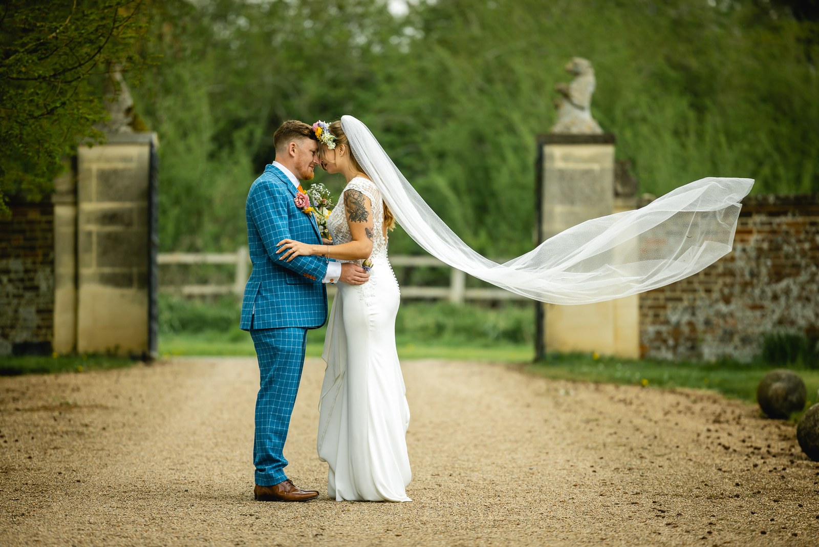 Bride and groom embracing on the drive between the gate piers, her veil flying