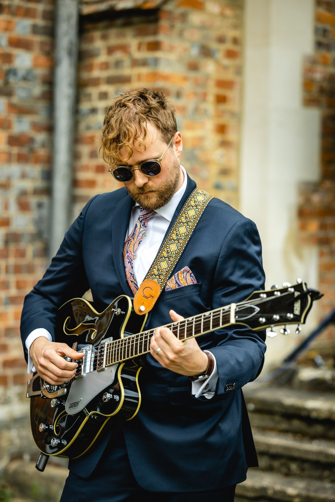 A guitarist in sunglasses playing a semi-acoustic guitar by a brick wall