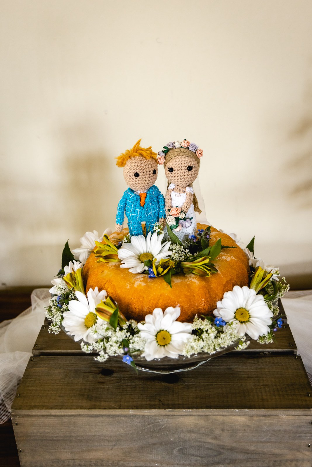 A knitted bride and groom cake topper on an orange bundt cake dressed with daisies