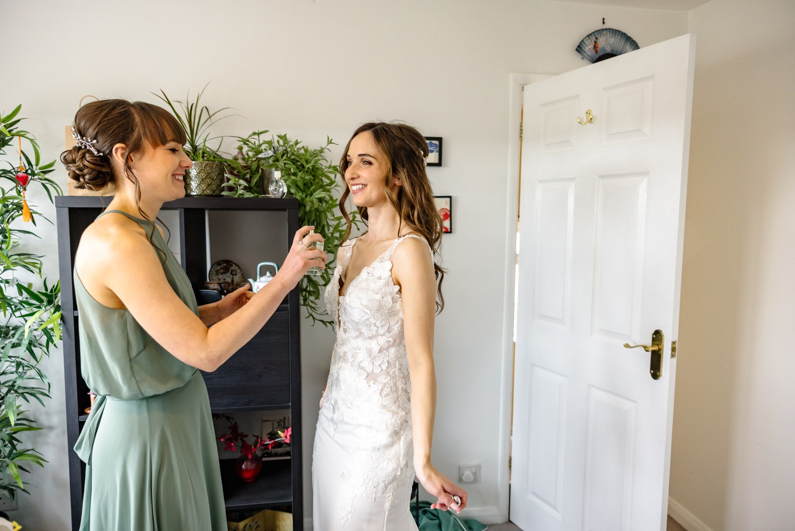 A bridesmaid in sage green helping the bride get ready by a shelf