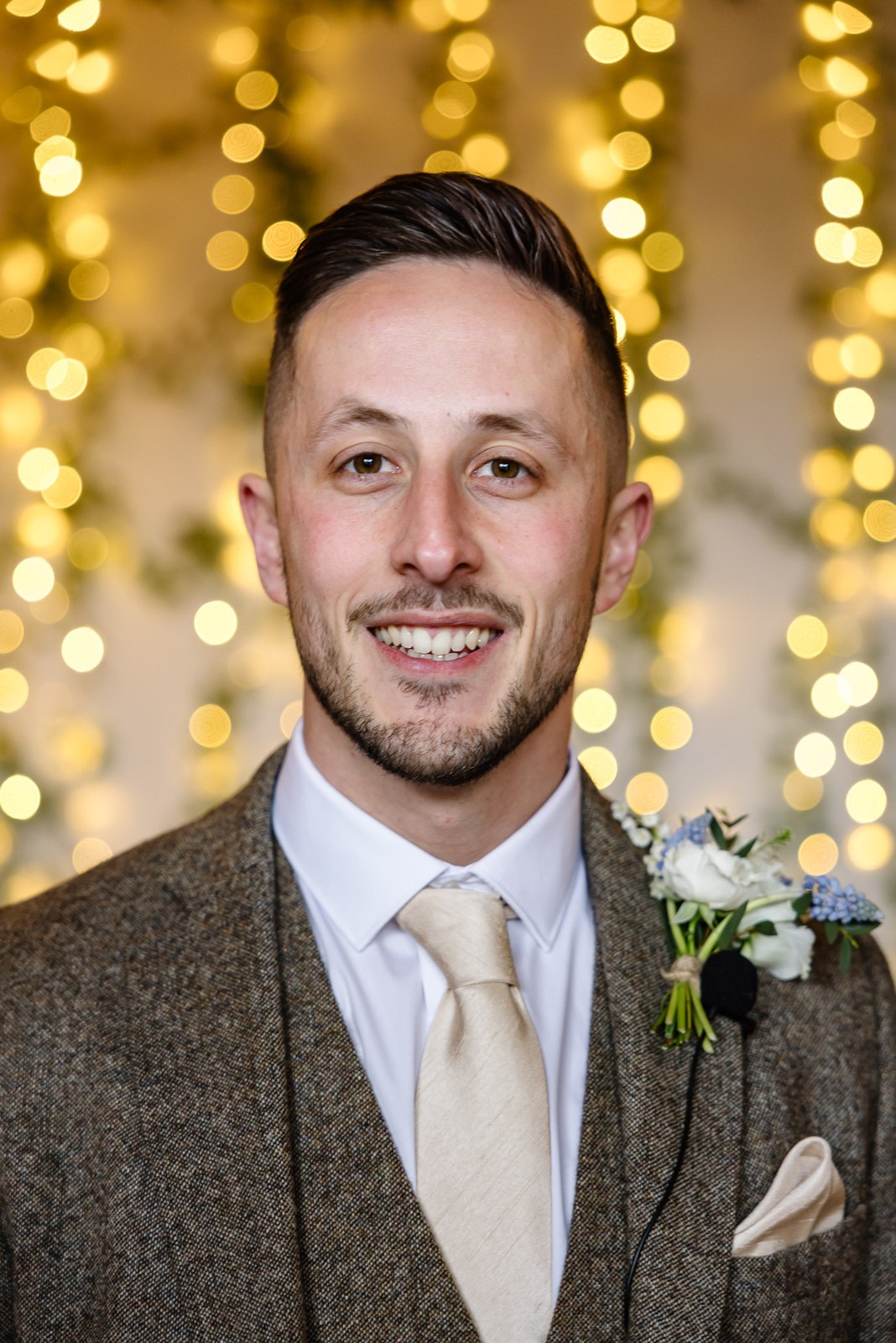 Portrait of the groom in a brown tweed suit against a fairy-light backdrop