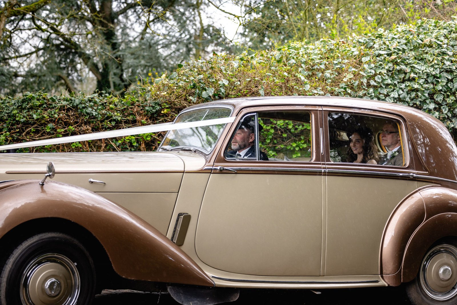 The bride arriving in a vintage cream and brown wedding car down a leafy lane