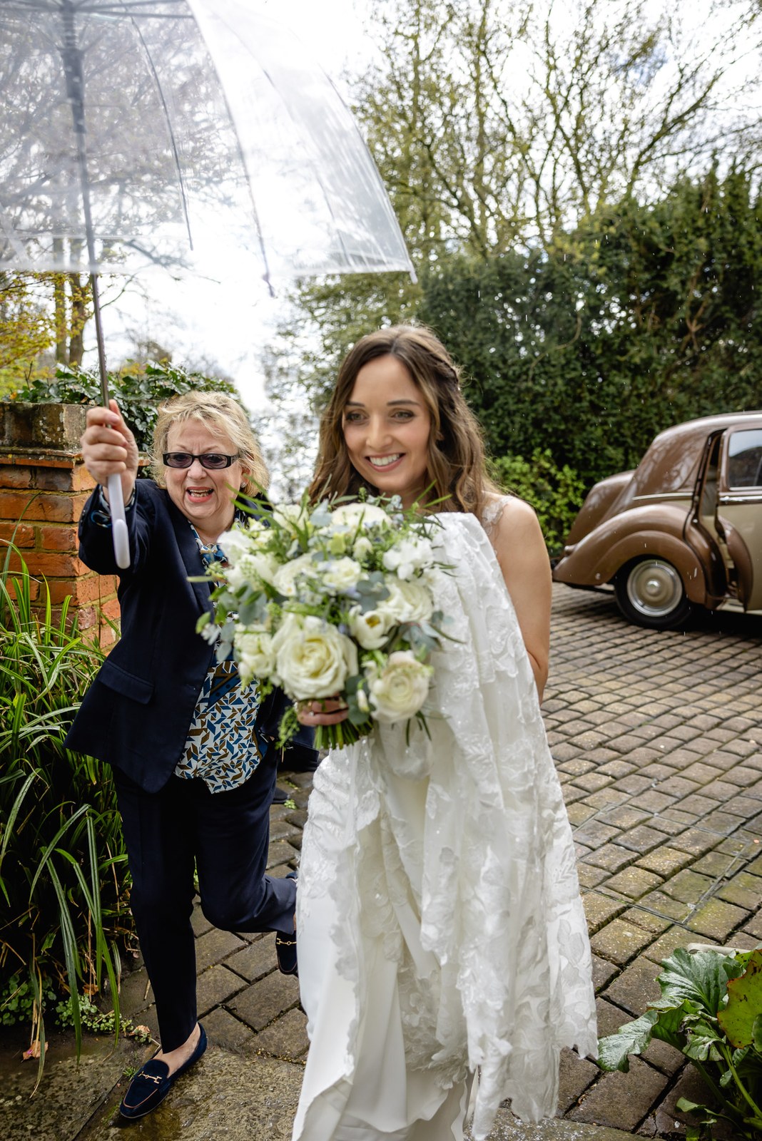 A guest holding a clear umbrella for the bride by the vintage car