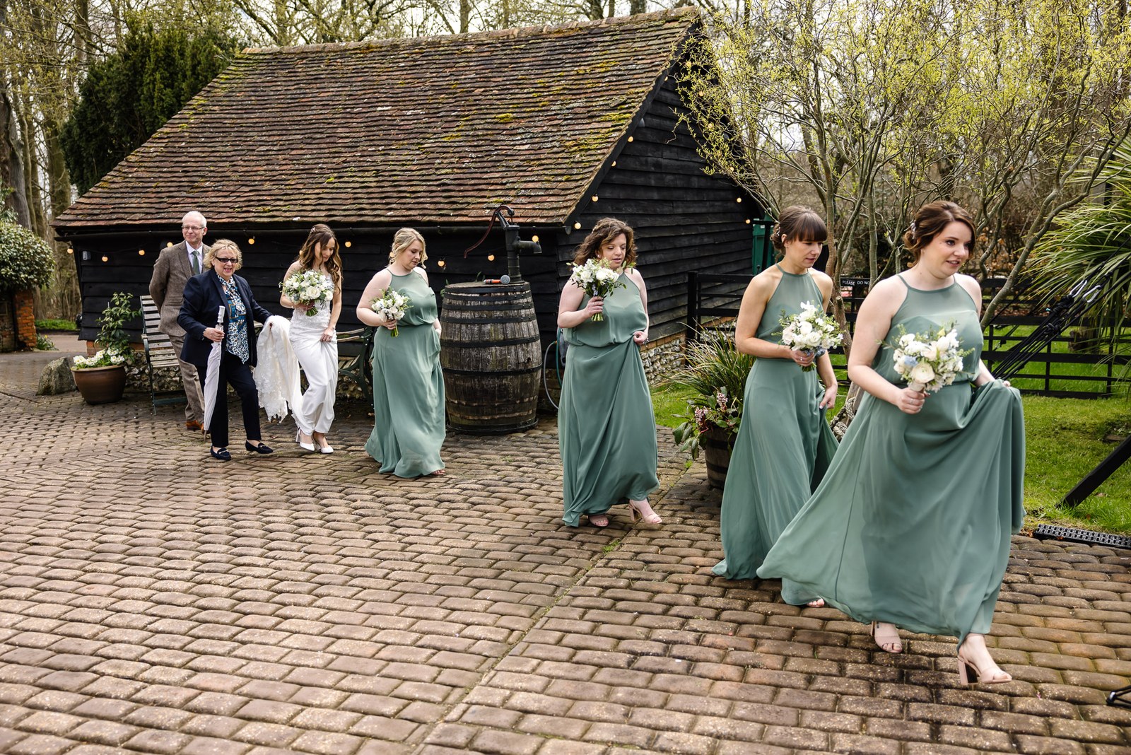 Bride, her father and sage-green bridesmaids crossing the courtyard at Old Luxters Barn