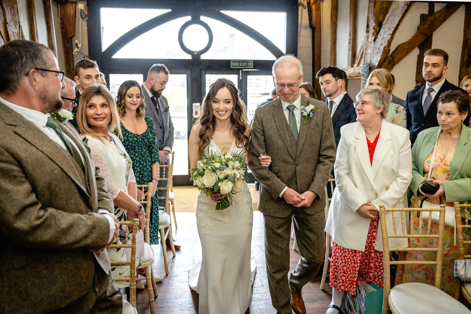 Bride entering on her father's arm through the arched glass doors of Old Luxters Barn