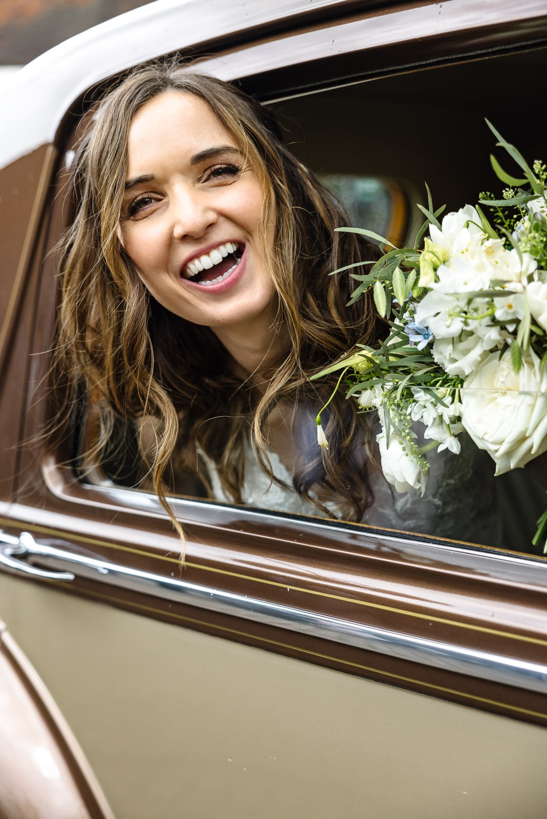 Bride laughing as she leans from the vintage car window with her bouquet