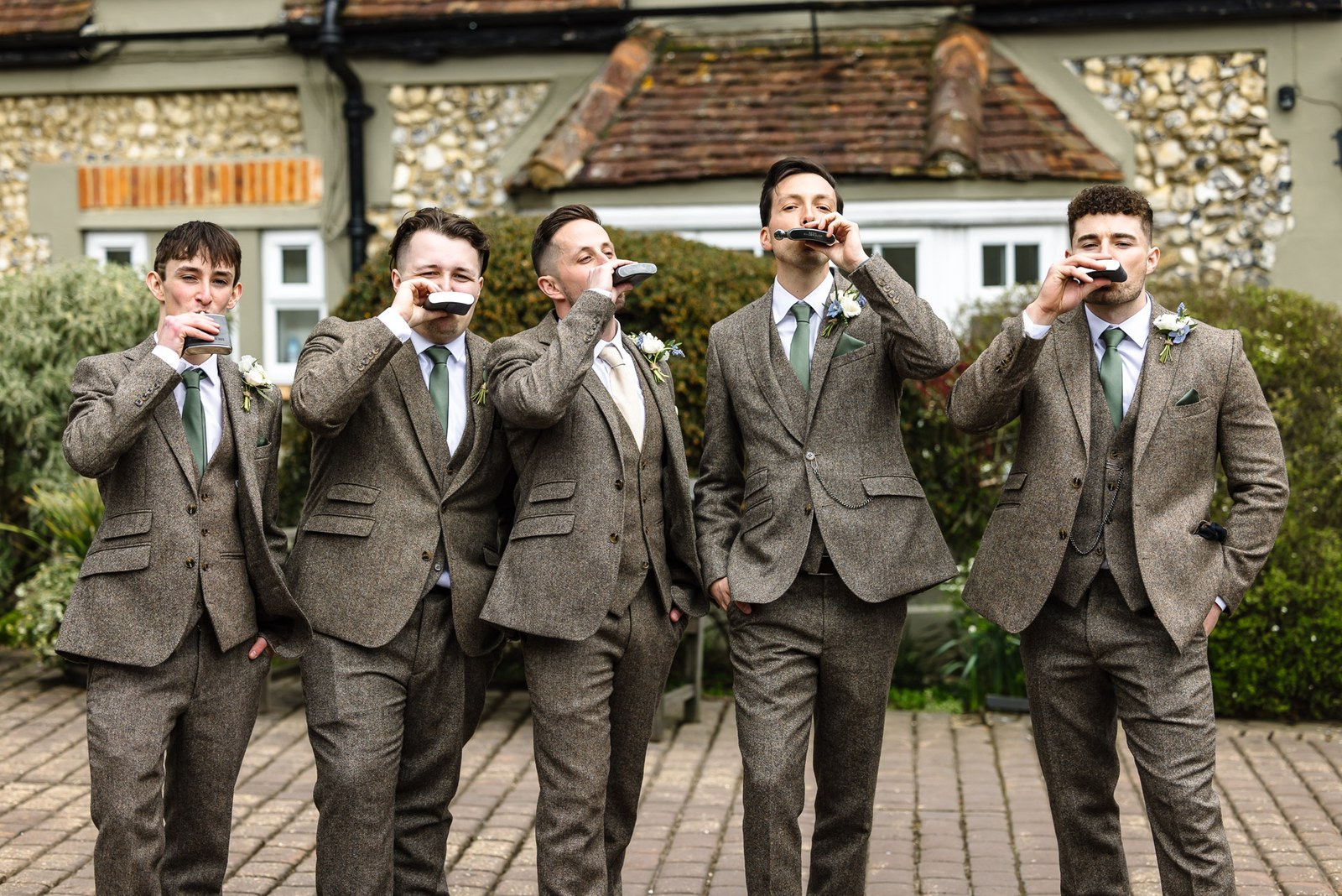 Groom and groomsmen in tweed downing drinks together in the courtyard, flint building behind