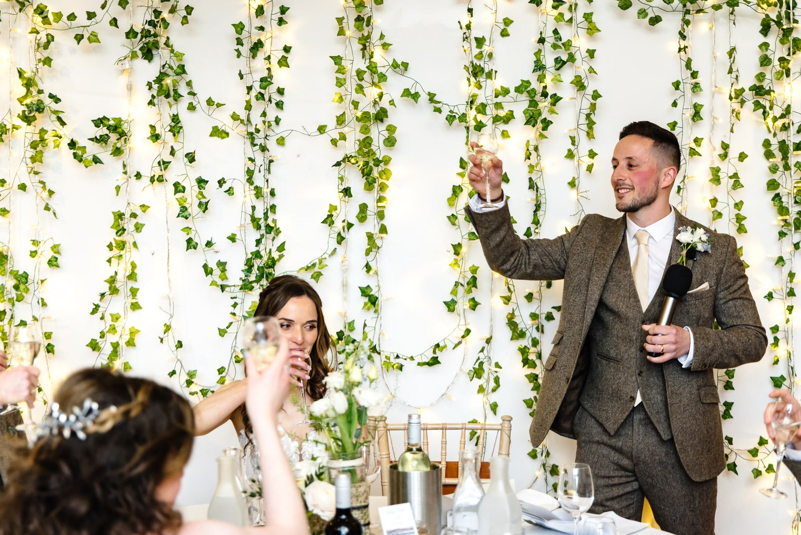 Groom raising a glass for his speech against an ivy and fairy-light backdrop