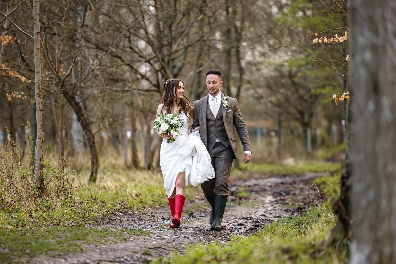 Bride and groom walking a muddy woodland track, the bride in red wellies