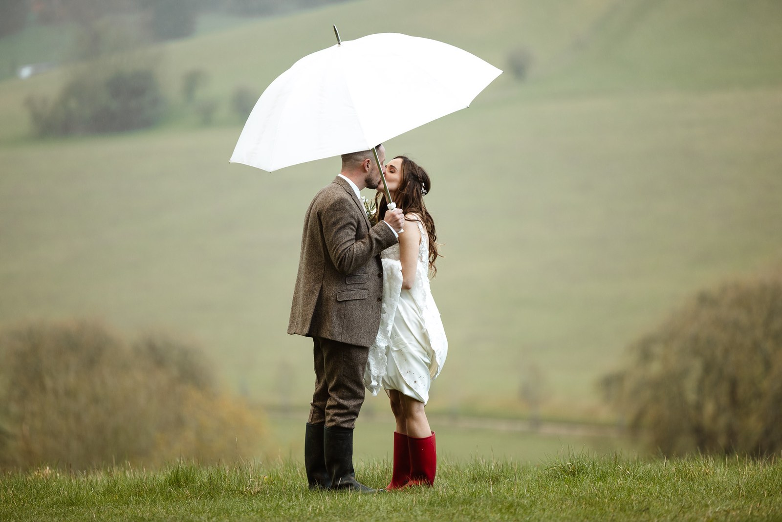 Bride and groom kissing under a white umbrella in a rolling green field