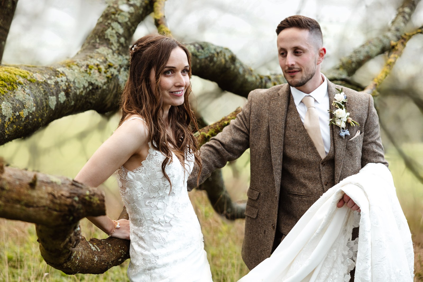 Bride and groom by a low tree branch in the meadow, the groom holding her train