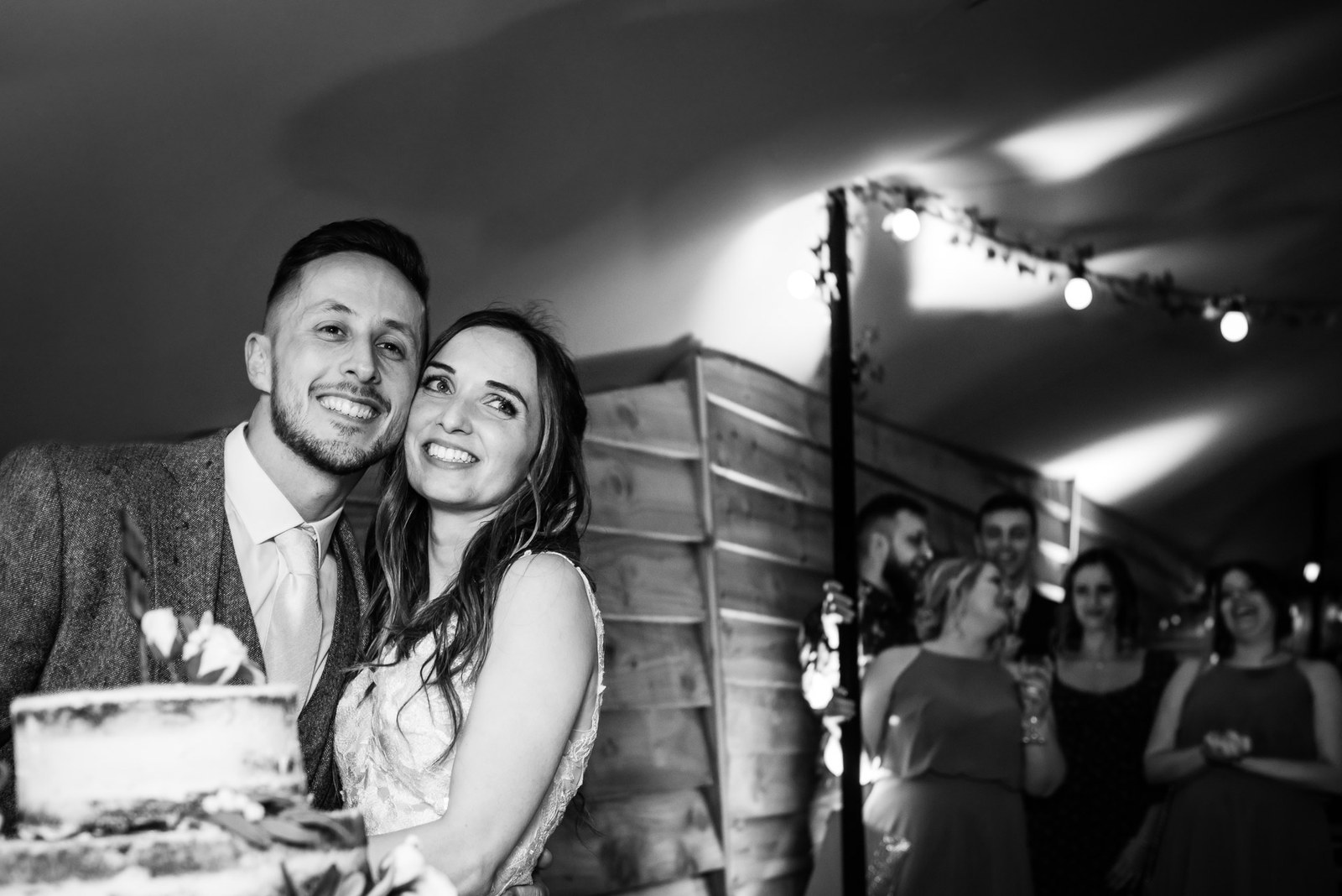 Black and white of the couple beside their cake under festoon lights