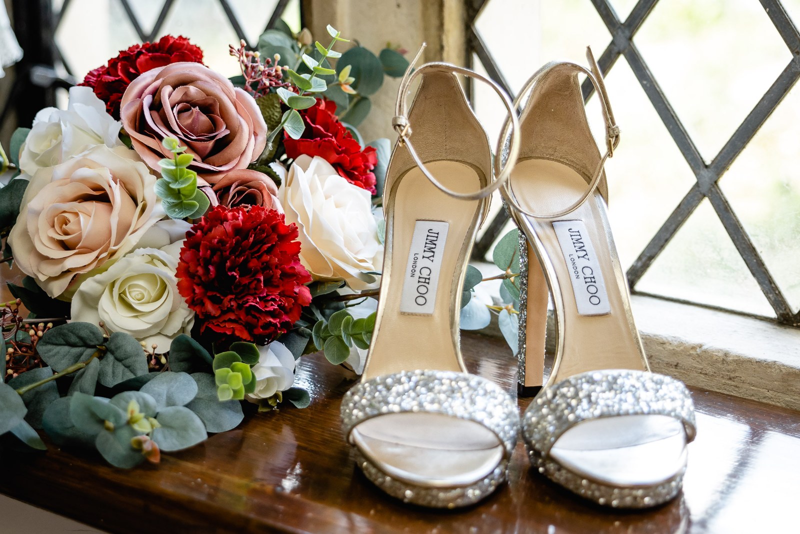 Sparkle-embellished bridal heels beside a red and cream rose bouquet on a windowsill