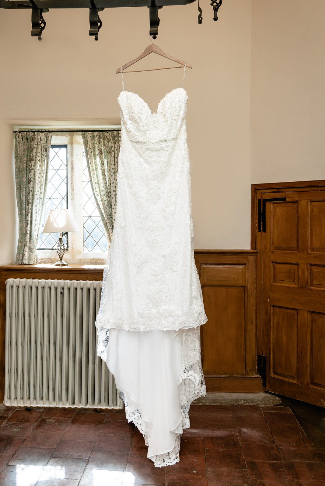 The bride's lace fishtail gown hanging in a wood-panelled room with a leaded window
