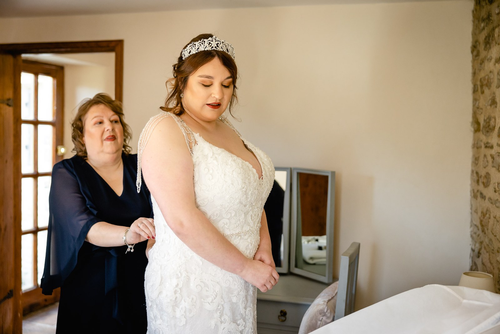 The bride's mother fastening the back of her lace wedding gown in the bridal suite