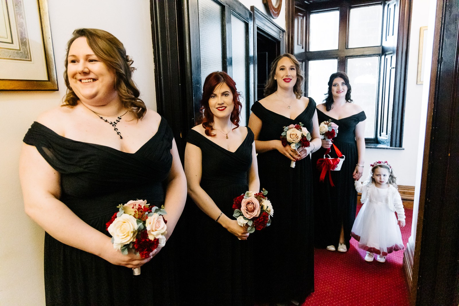 Bridesmaids in black gowns lined up with a flower girl on a red carpet
