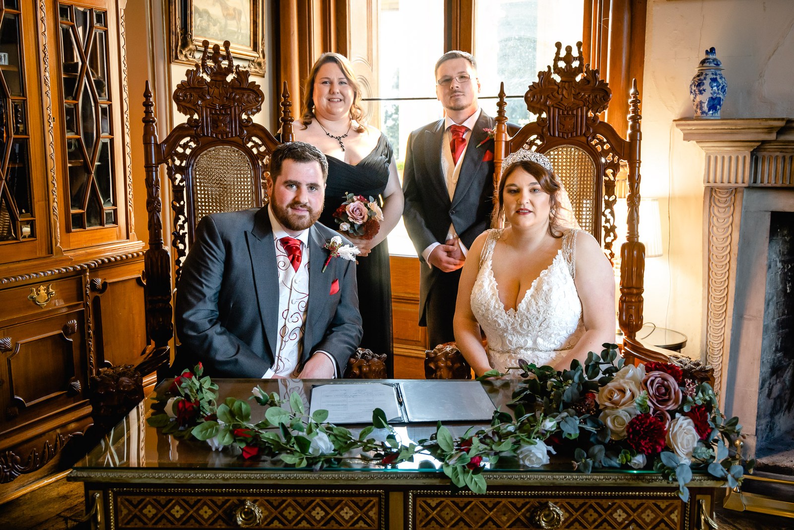The couple seated in ornate carved chairs to sign the register amid floral displays