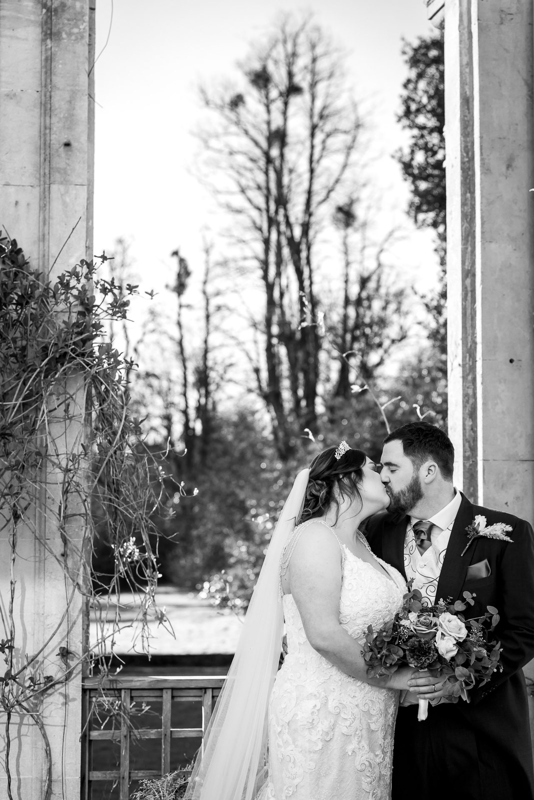 Black-and-white portrait of the couple kissing framed by tall stone columns