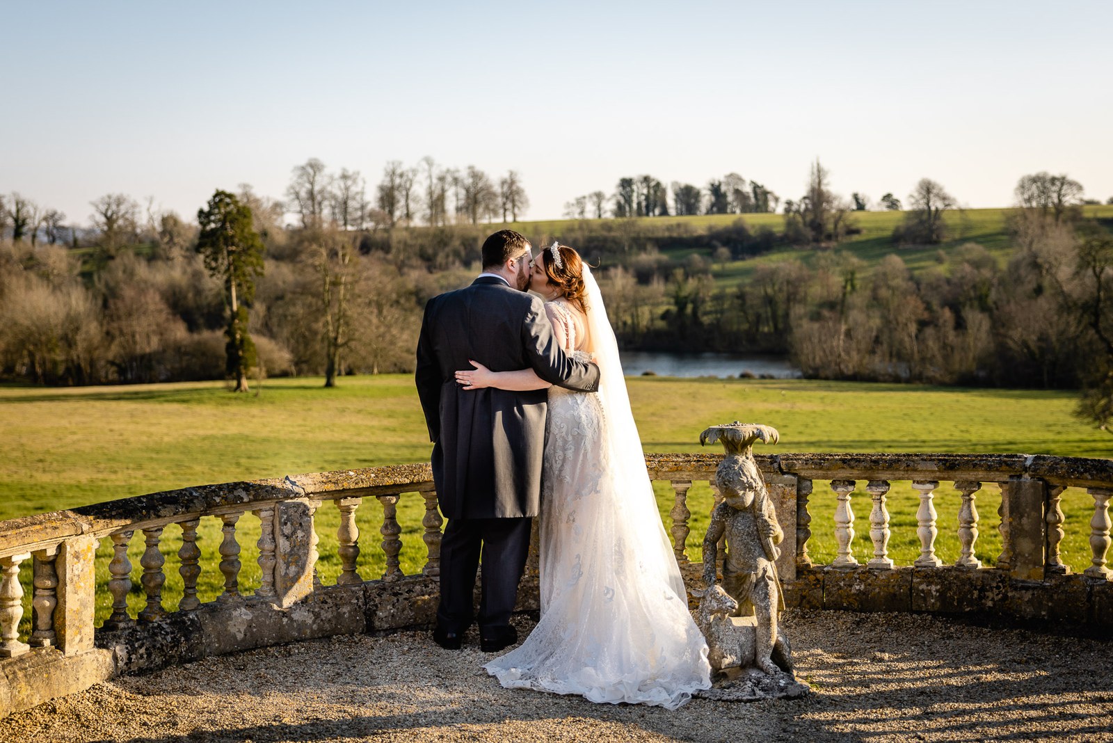 The couple embracing on a stone balustrade above the parkland and lake at Orchardleigh House
