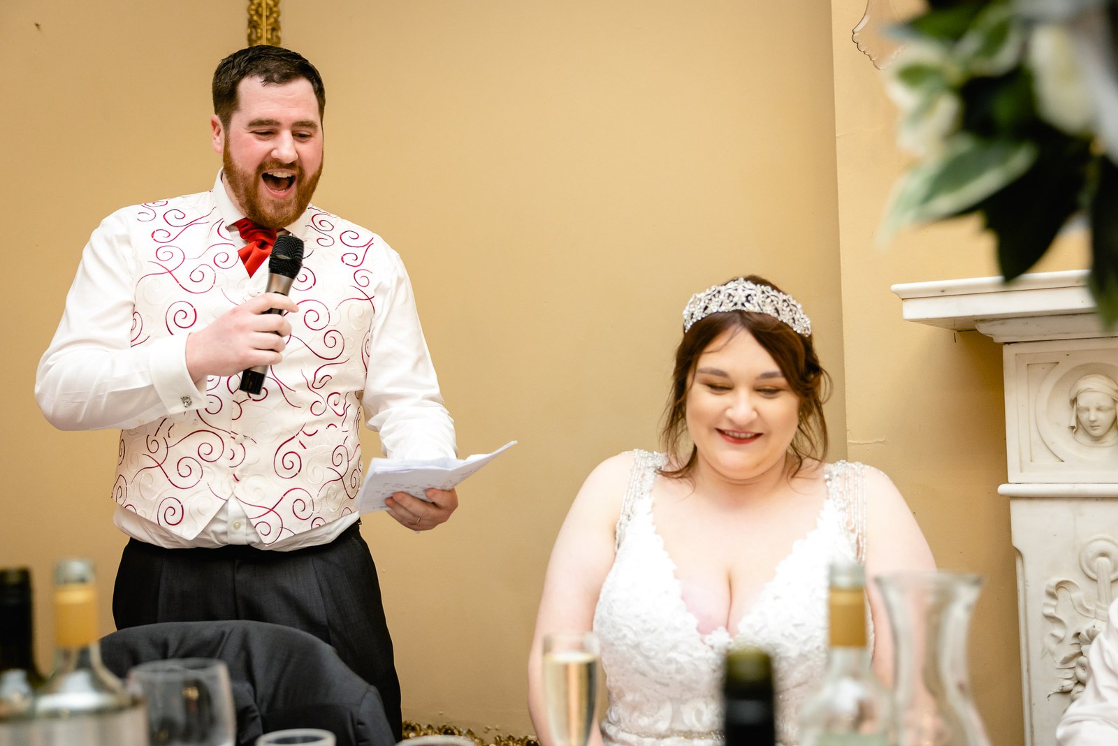 The groom giving his speech with microphone and notes as the bride laughs beside him