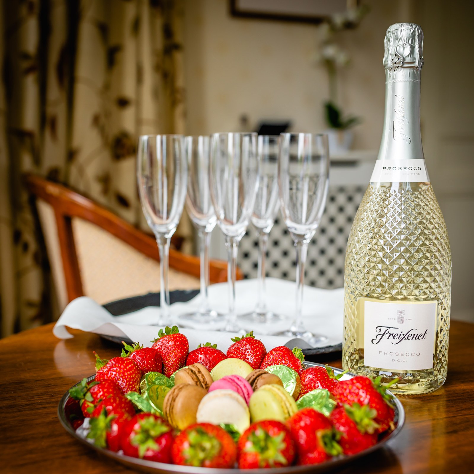 A bottle of prosecco with champagne flutes, strawberries and macarons on a table