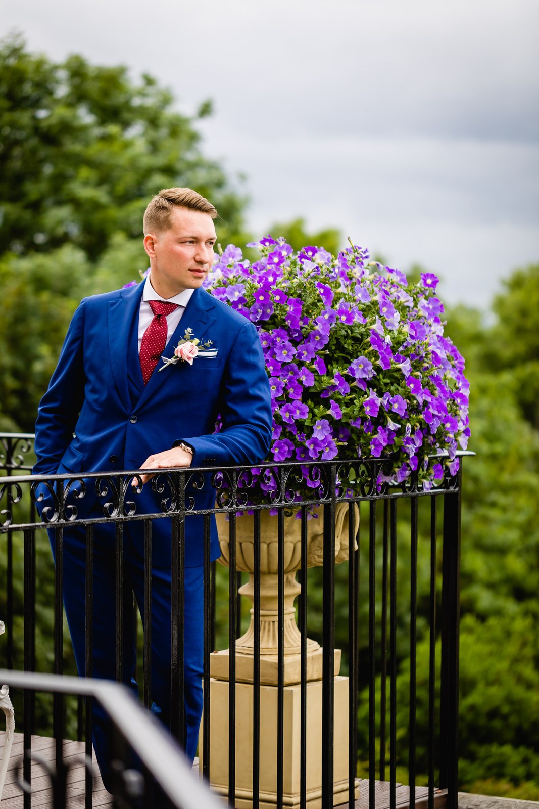 The groom leaning on a balcony railing beside purple petunias at The Petersham Hotel