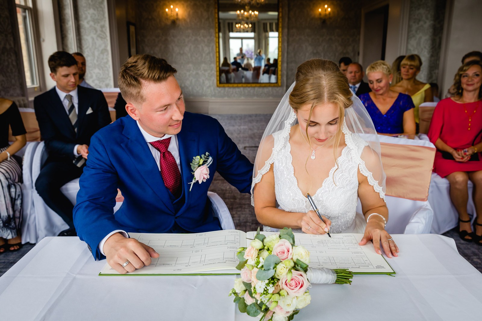 The couple signing the register at a flower-topped table as guests watch
