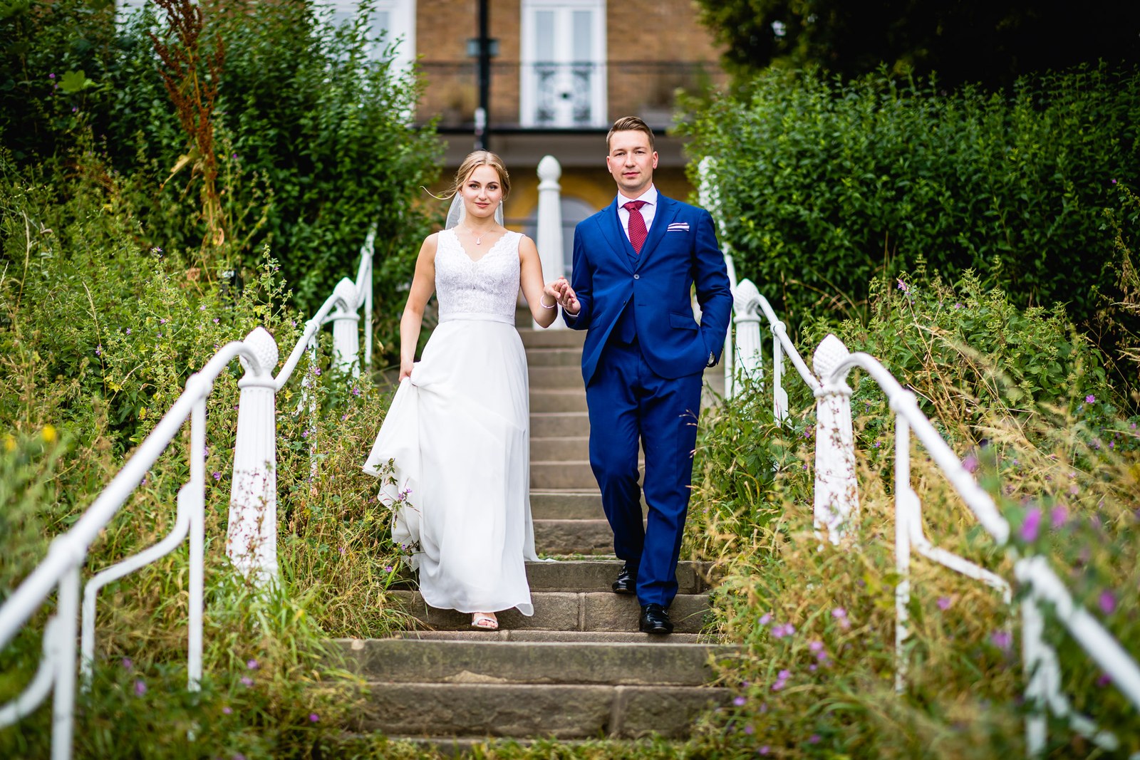 The couple walking down white-railed stone steps between overgrown greenery