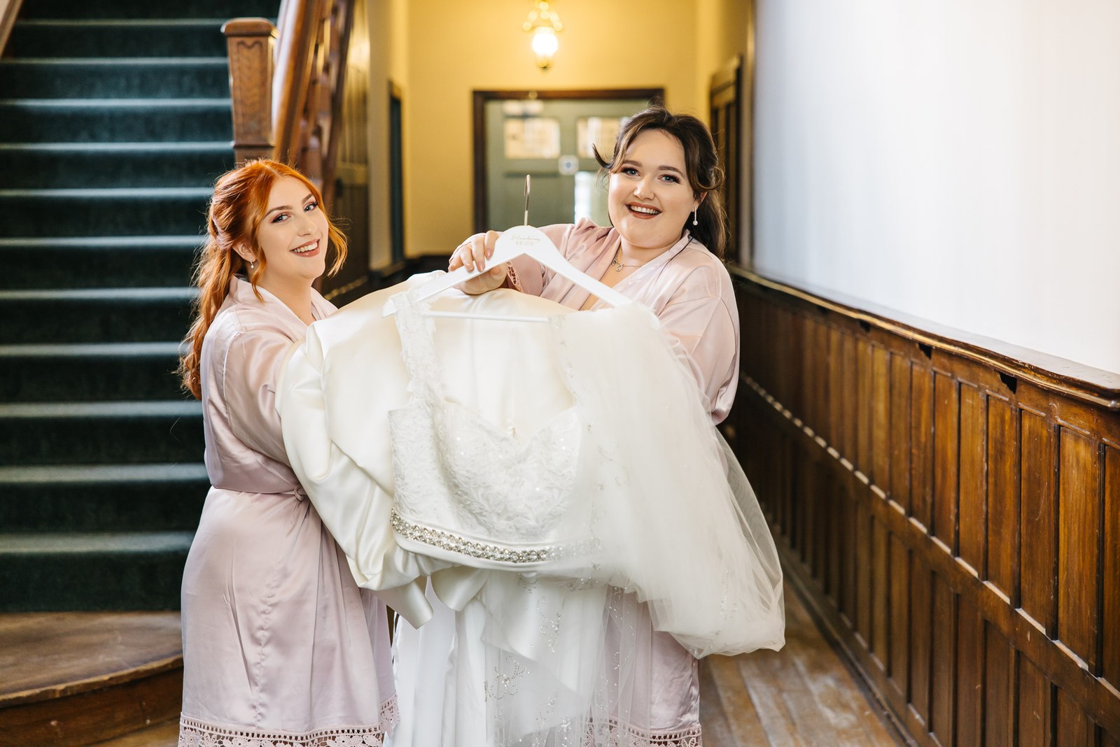 Two bridesmaids in robes carrying the bride's gown in a wood-panelled hallway