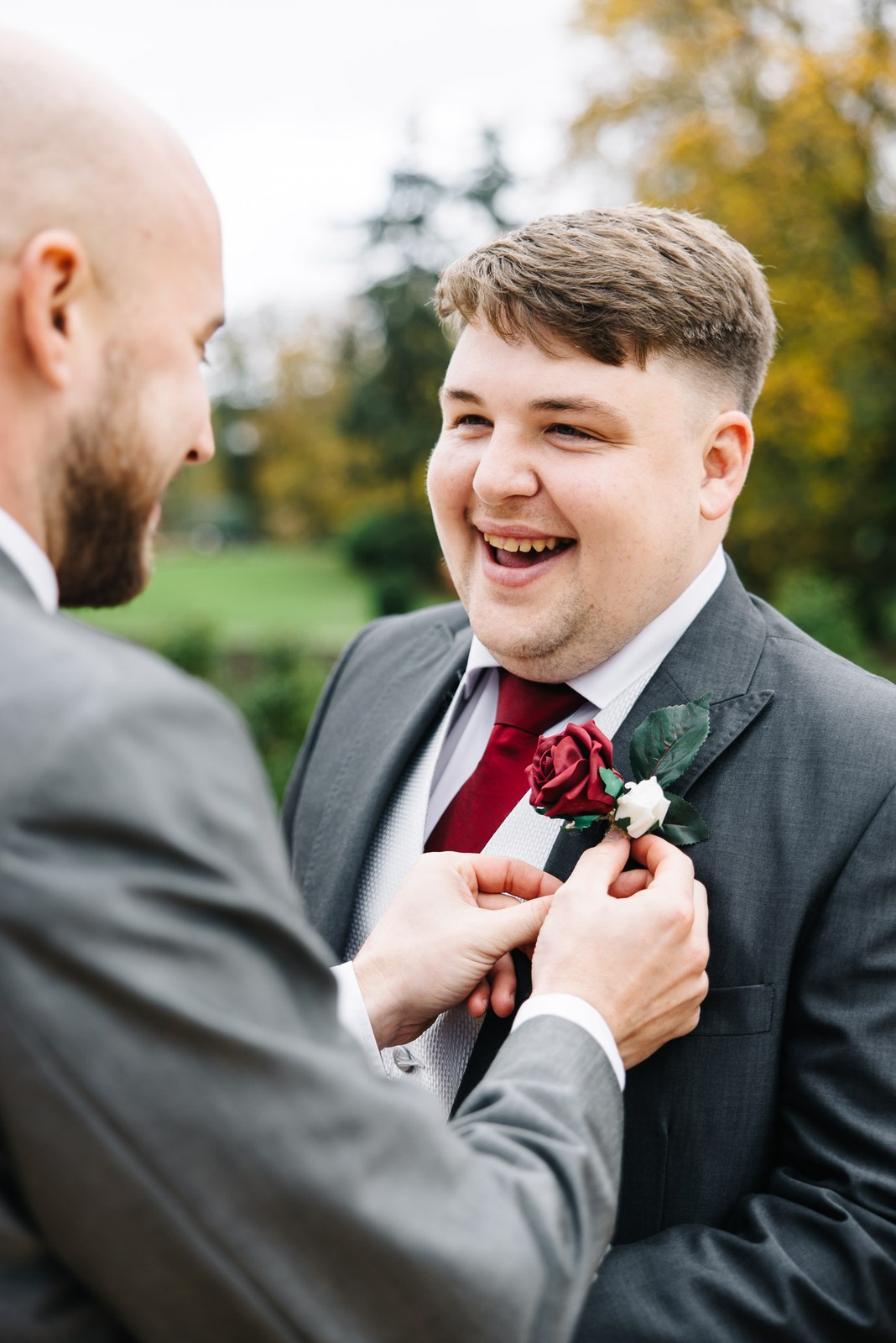 A man in grey pinning the groom's buttonhole outdoors amid autumn trees