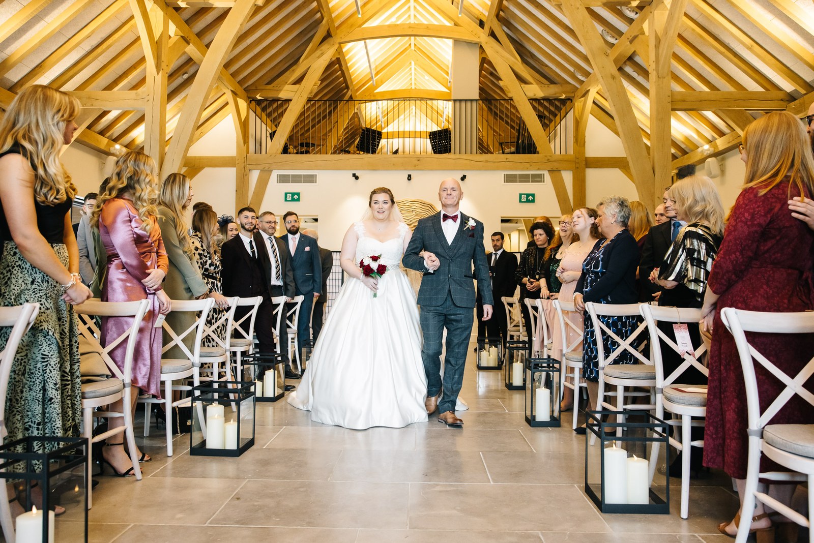 The bride walking down the aisle on her father's arm beneath the oak beams at The Post Barn