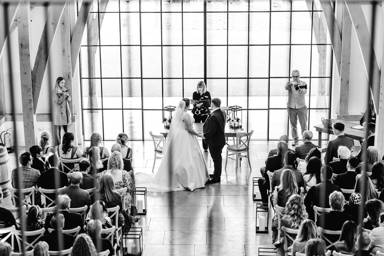 Black-and-white wide shot of the ceremony before the tall windows at The Post Barn