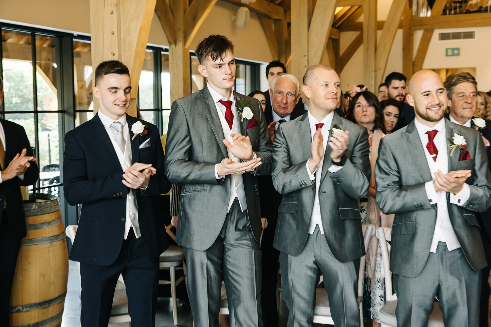 Guests and groomsmen applauding beside wine barrels under the oak beams at The Post Barn