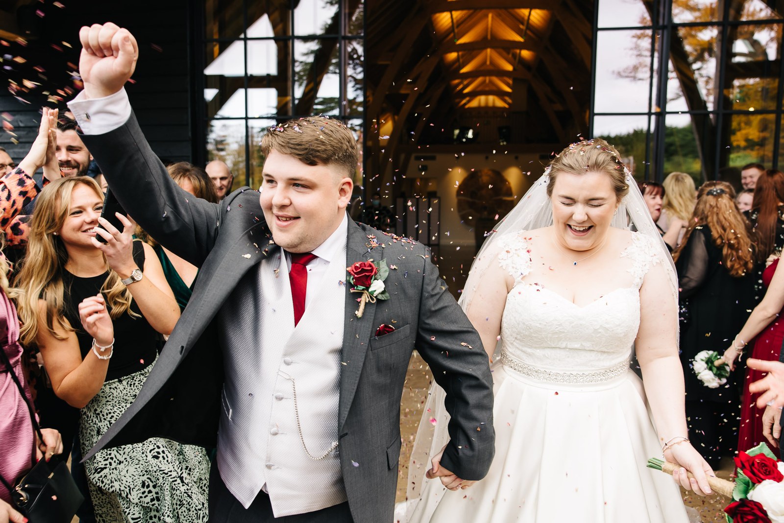 The couple stepping through confetti with arms raised beneath the barn beams
