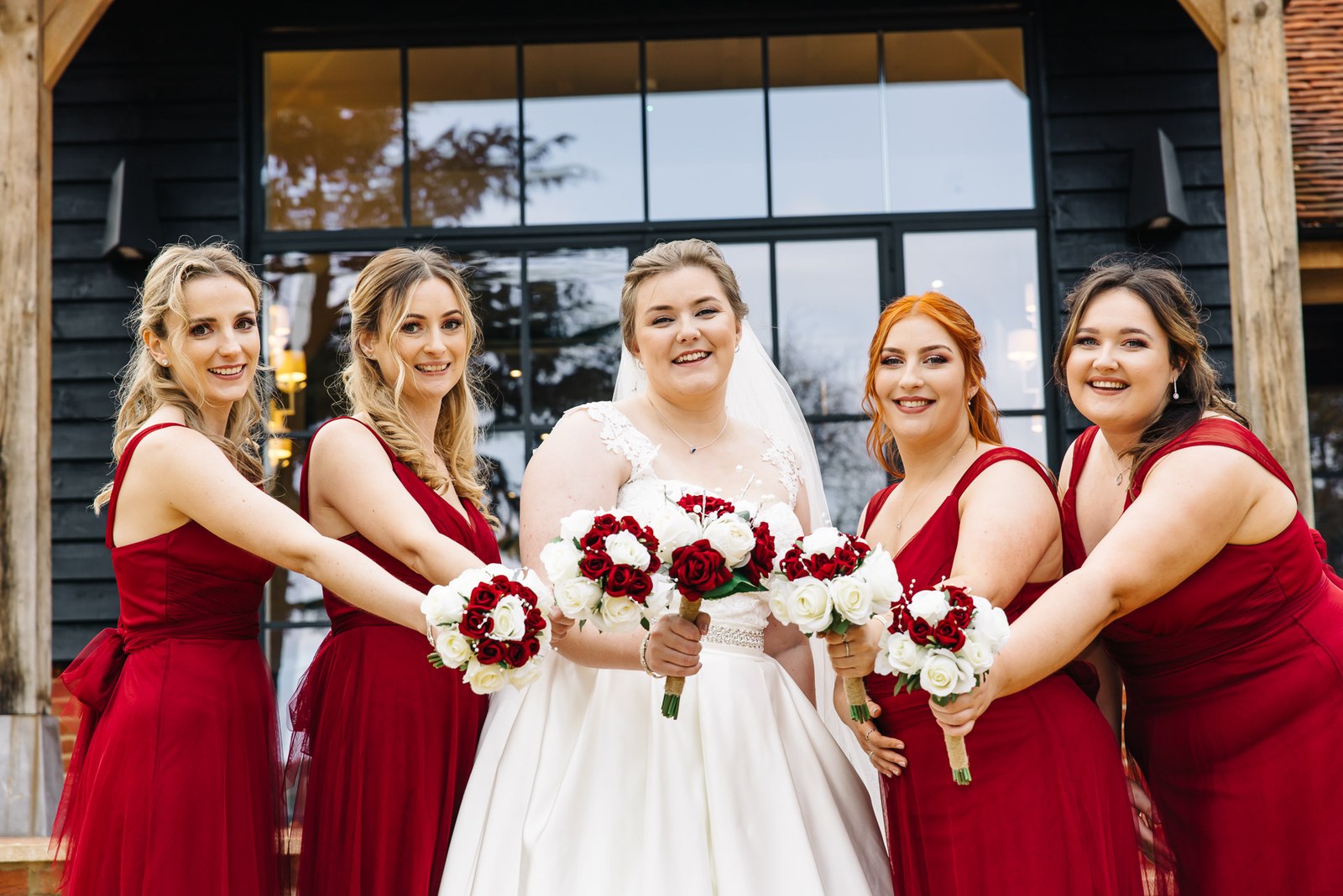 The bride and bridesmaids in red holding out their bouquets before the venue's tall windows