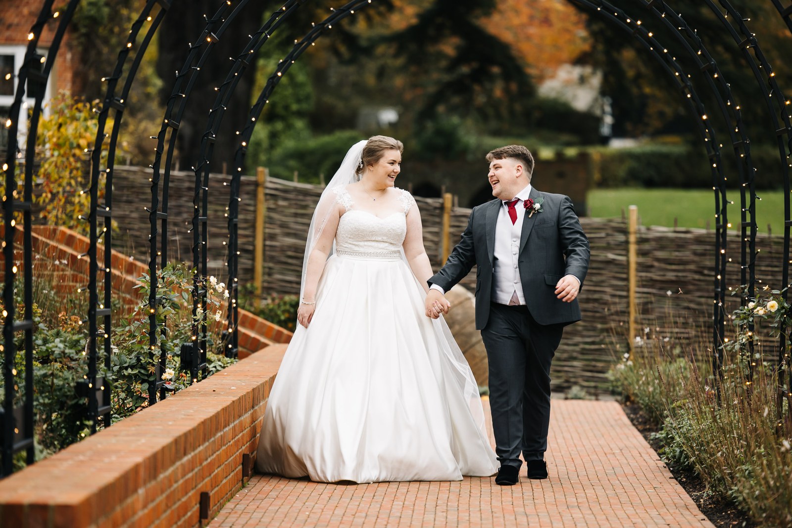 The couple walking hand in hand beneath a fairy-lit arch in the gardens at The Post Barn