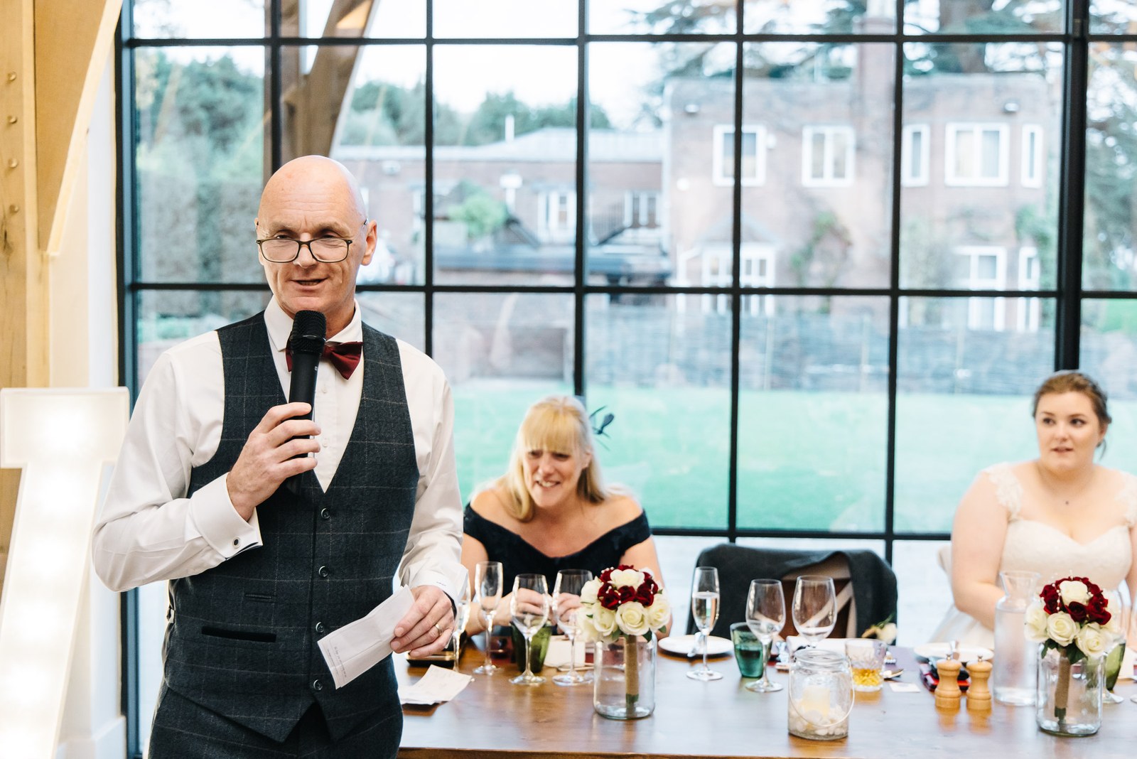 A father giving his speech at the top table before the tall windows of The Post Barn