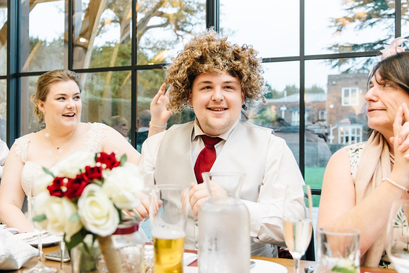 The bride laughing as the groom tries on a curly wig during the speeches
