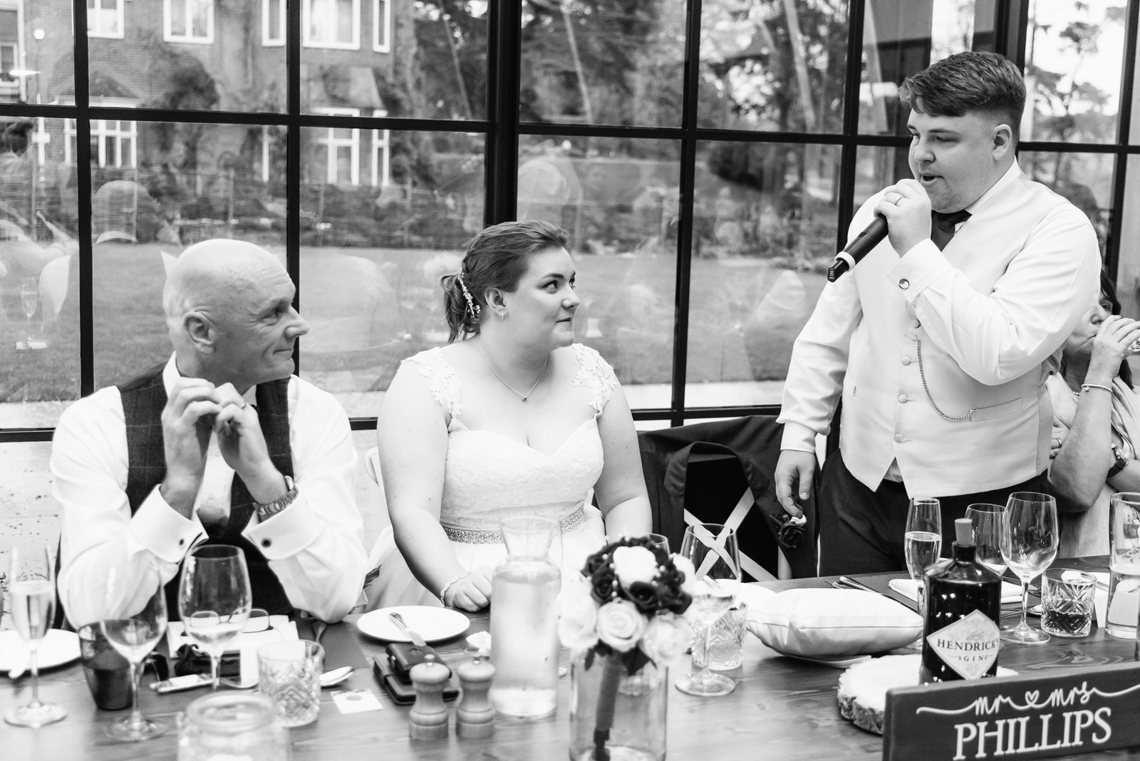 Black-and-white shot of the groom speaking beside the personalised couple's sign