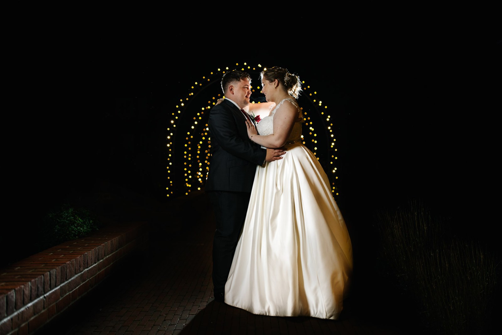 The couple embracing at night lit by a glowing fairy-light arch