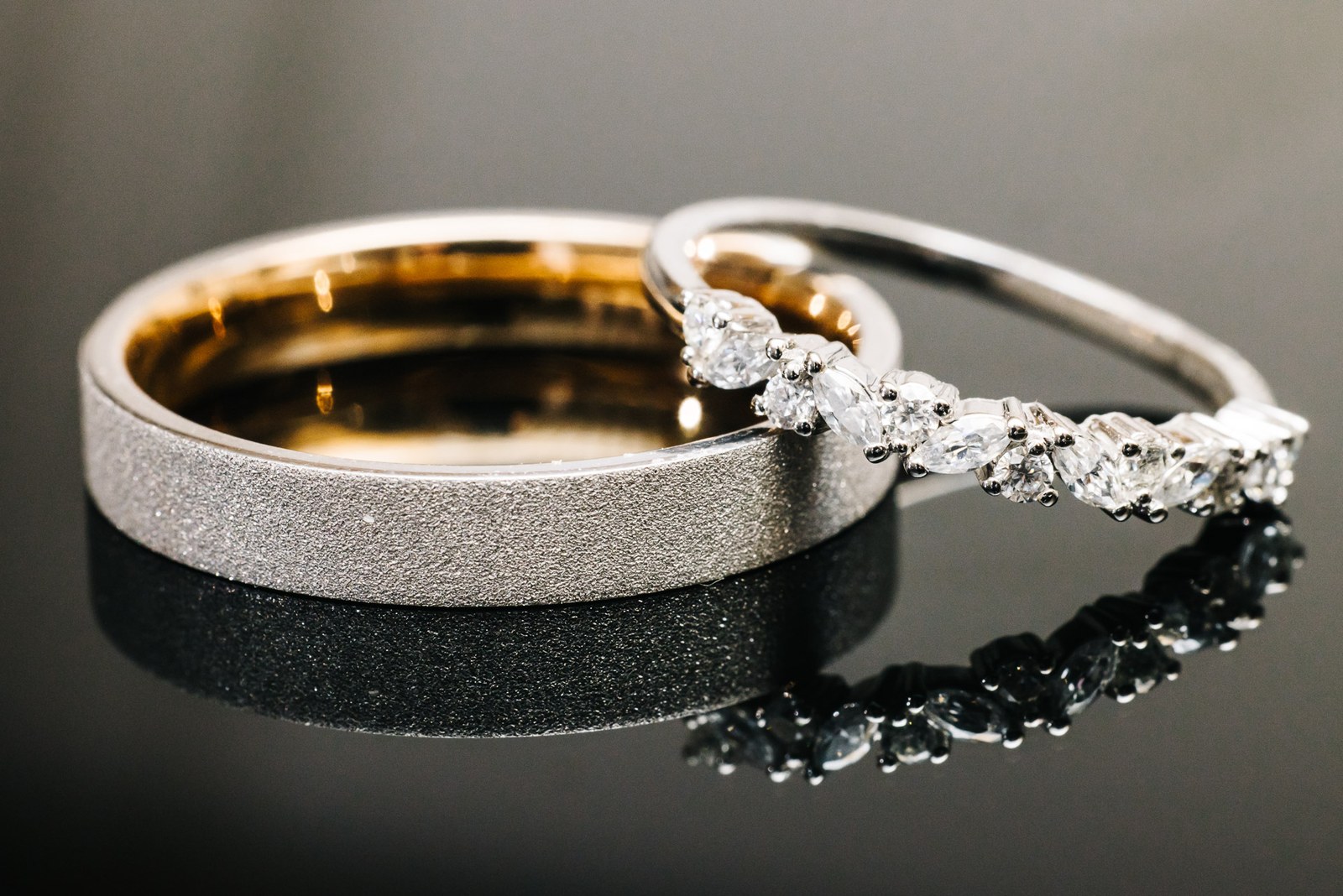 The two wedding rings, a textured band and a diamond band, on a dark reflective surface