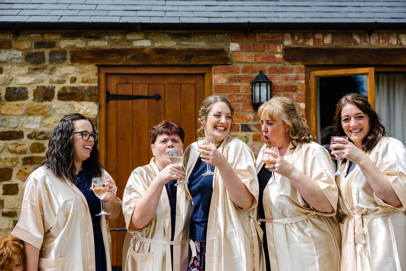 Bridesmaids and a bride in champagne satin robes toast outside the stone barn at Slapton Manor